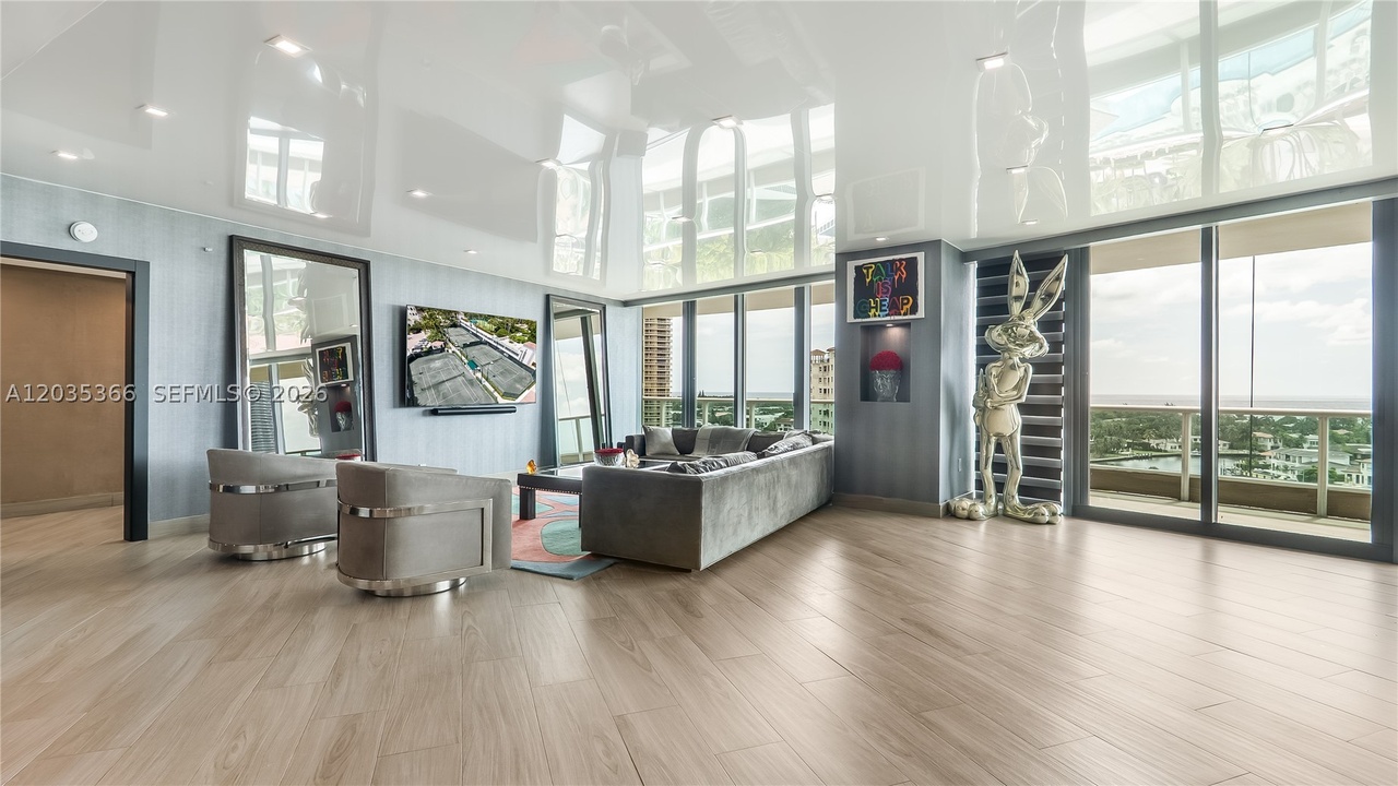 The double-height great room with the glossy white reflective ceiling, a wall of glass on two sides, gray seating, and a chrome sculpture by the windows