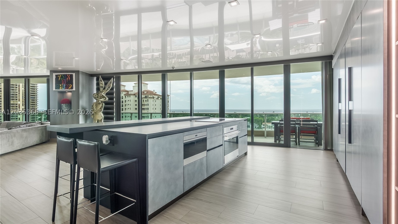 The Mia Cucina kitchen with a long waterfall island, integrated Wolf wall ovens, gray cabinetry, and the glass wall and terrace beyond