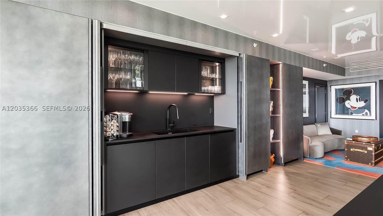 A concealed wet bar with matte charcoal cabinetry, backlit glass-front cabinets holding stemware, a bar sink, and pocketing doors that close it away