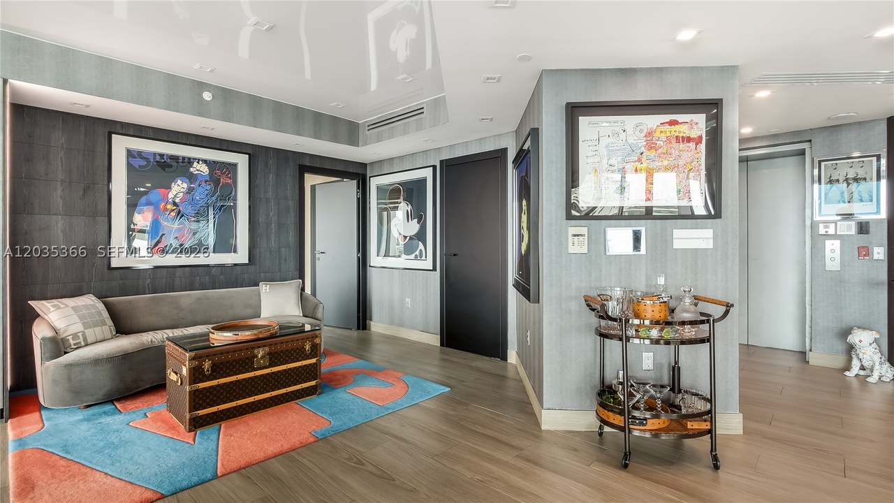 An interior sitting room beneath the glossy reflective ceiling, with a curved gray settee, a monogrammed steamer trunk used as a table, a bar cart, and framed pop-art across the gray-paneled walls