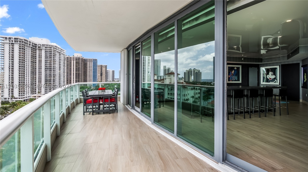 The wraparound terrace running along the curved glass wall of the residence, with a dining set, a glass-and-steel railing, and the Sunny Isles skyline and ocean beyond