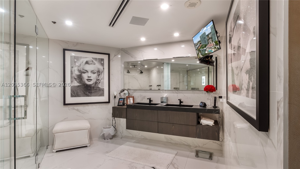The primary bath in book-matched white marble with a floating dark-wood double vanity, a wide mirror, a framed Marilyn Monroe print, and a glass shower at left