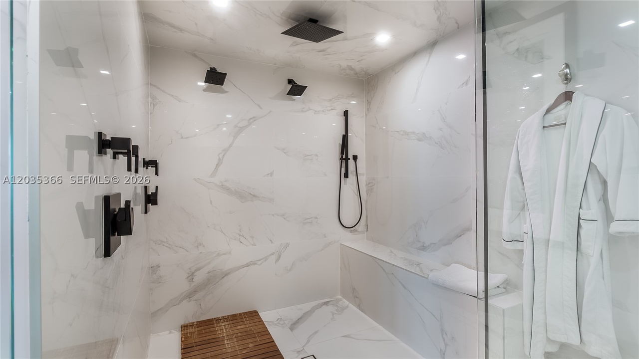 The walk-in marble shower with multiple rain and wall jets in matte black, a marble bench, a teak floor mat, and a robe hanging at the glass
