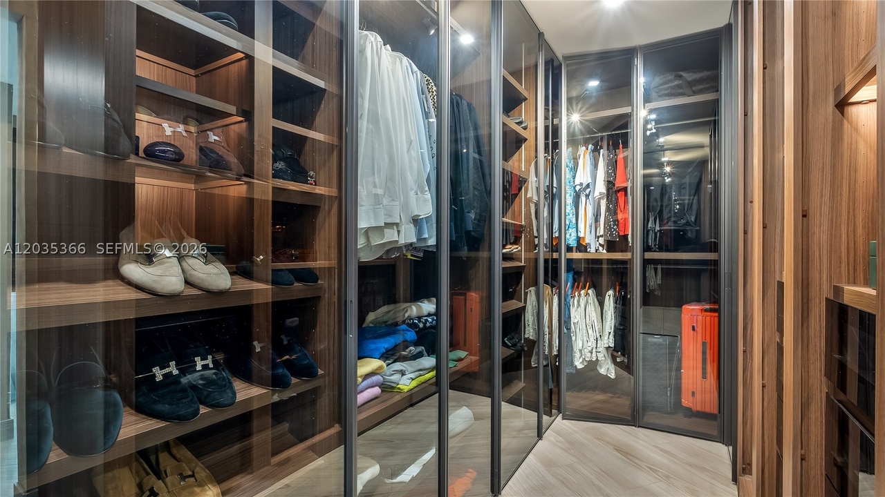 A walk-through dressing room lined with glass-front wood cabinetry, lit shelving displaying shoes and bags, and hanging wardrobe on both sides