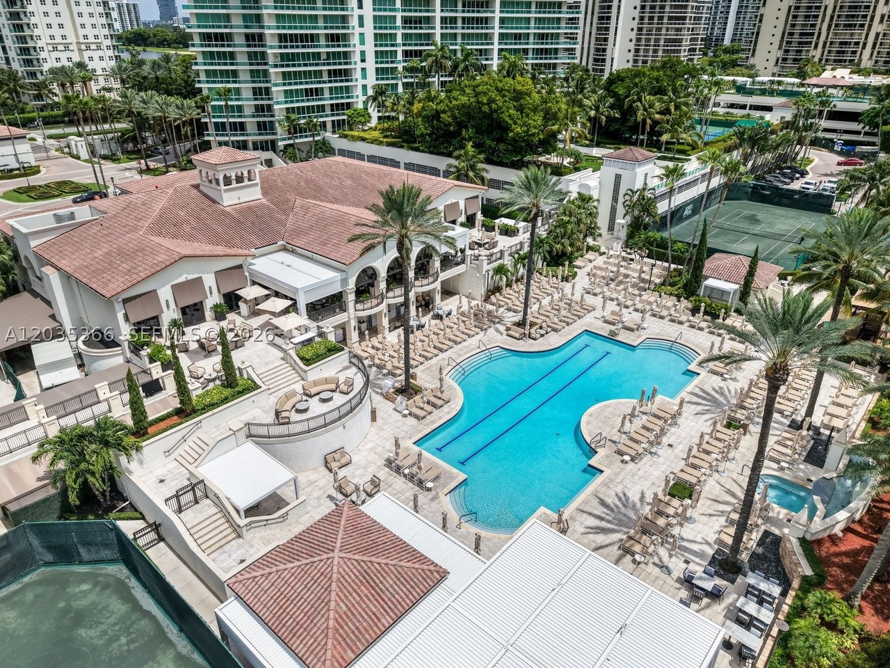 An aerial of the Porto Vita resort pool and clubhouse, with lounge chairs ringing the lap pool, cabanas, palms, and tennis courts behind
