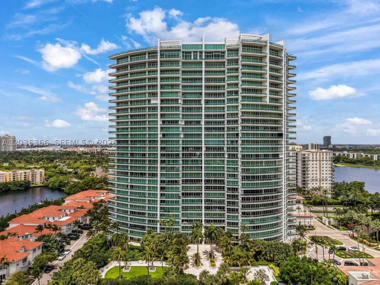The curved green-glass Porto Vita tower seen from across the lake, rising above a low row of red-roofed villas and palms